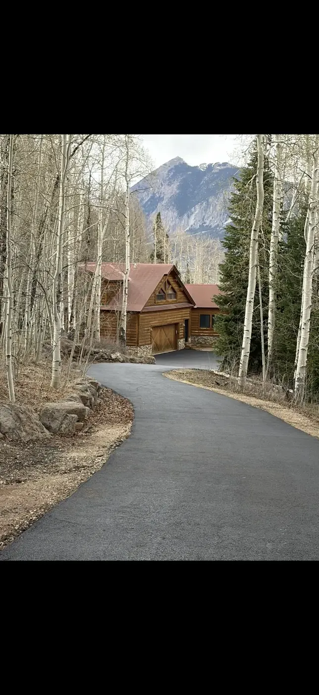 Asphalt driveway winding through aspens to a log cabin below a peak
